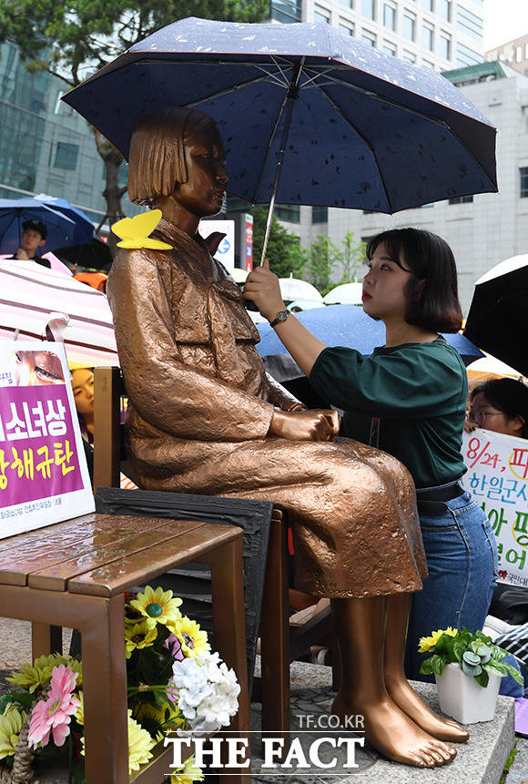 제 1397차 일본군 성노예제 문제 해결을 위한 정기 수요집회가 24일 오후 서울 종로구 옛 일본대사관 앞에서 열린 가운데 한 참가자가 갑작스레 내리는 비를 피하기 위해 소녀상과 함께 우산을 쓰고 있다. /남용희 기자
